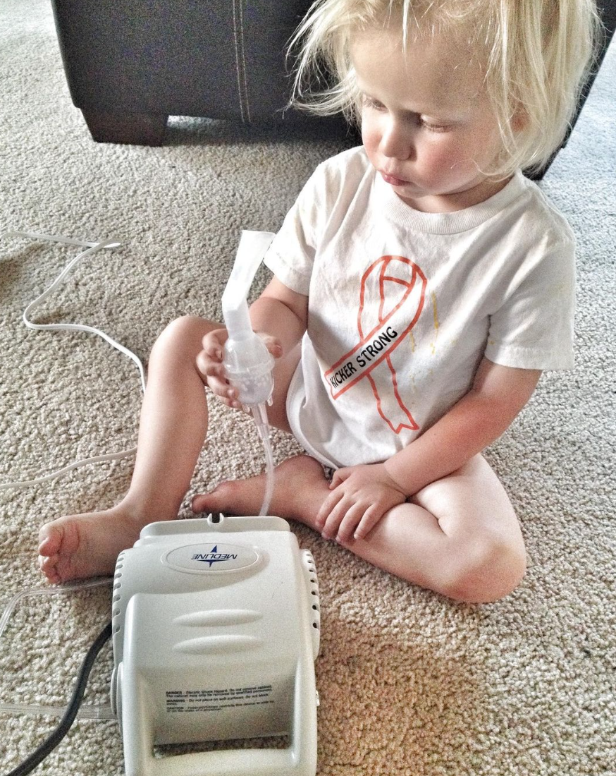 small child with cancer nebulizing wearing a white shirt with a cancer ribbon