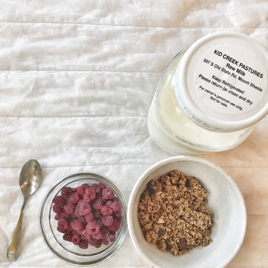 a white table cloth with a bowl of granola and raspberries and a jar of raw milk next to it.