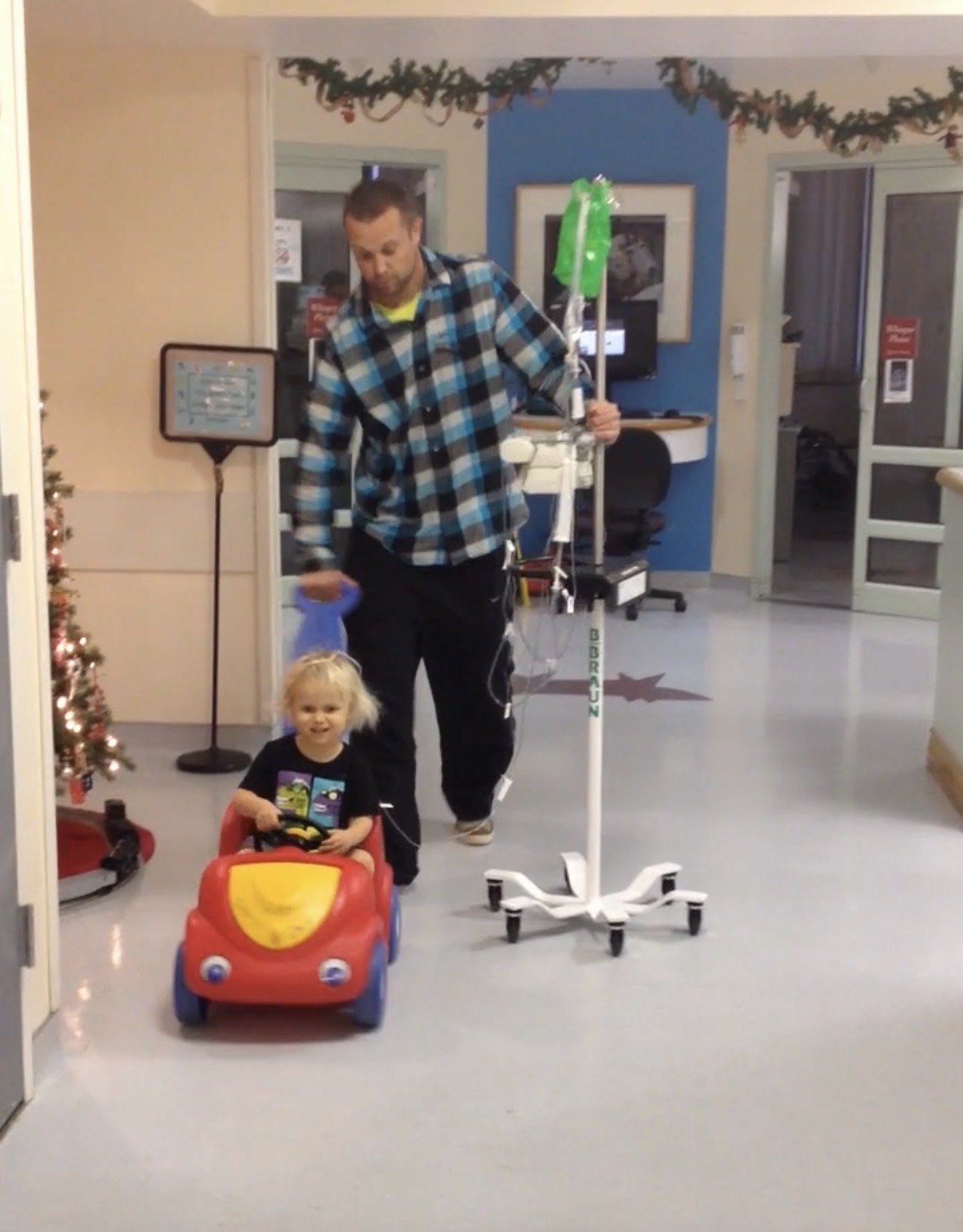 dad pushing a child through the hospital in a red car with Christmas decorations in the hall 