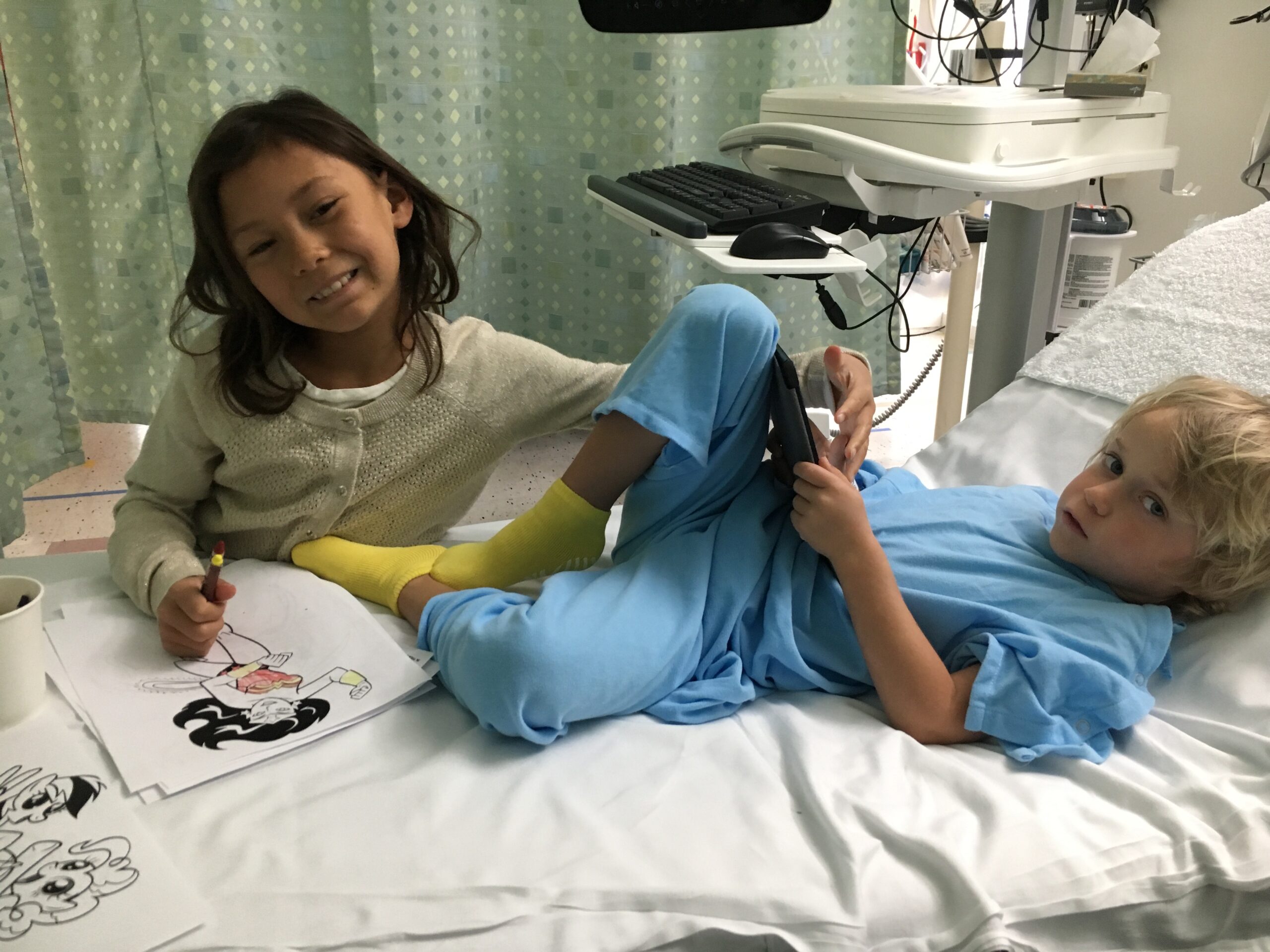 little sister and brother in the hospital, brother wearing a hospital gown, sister siting next to him