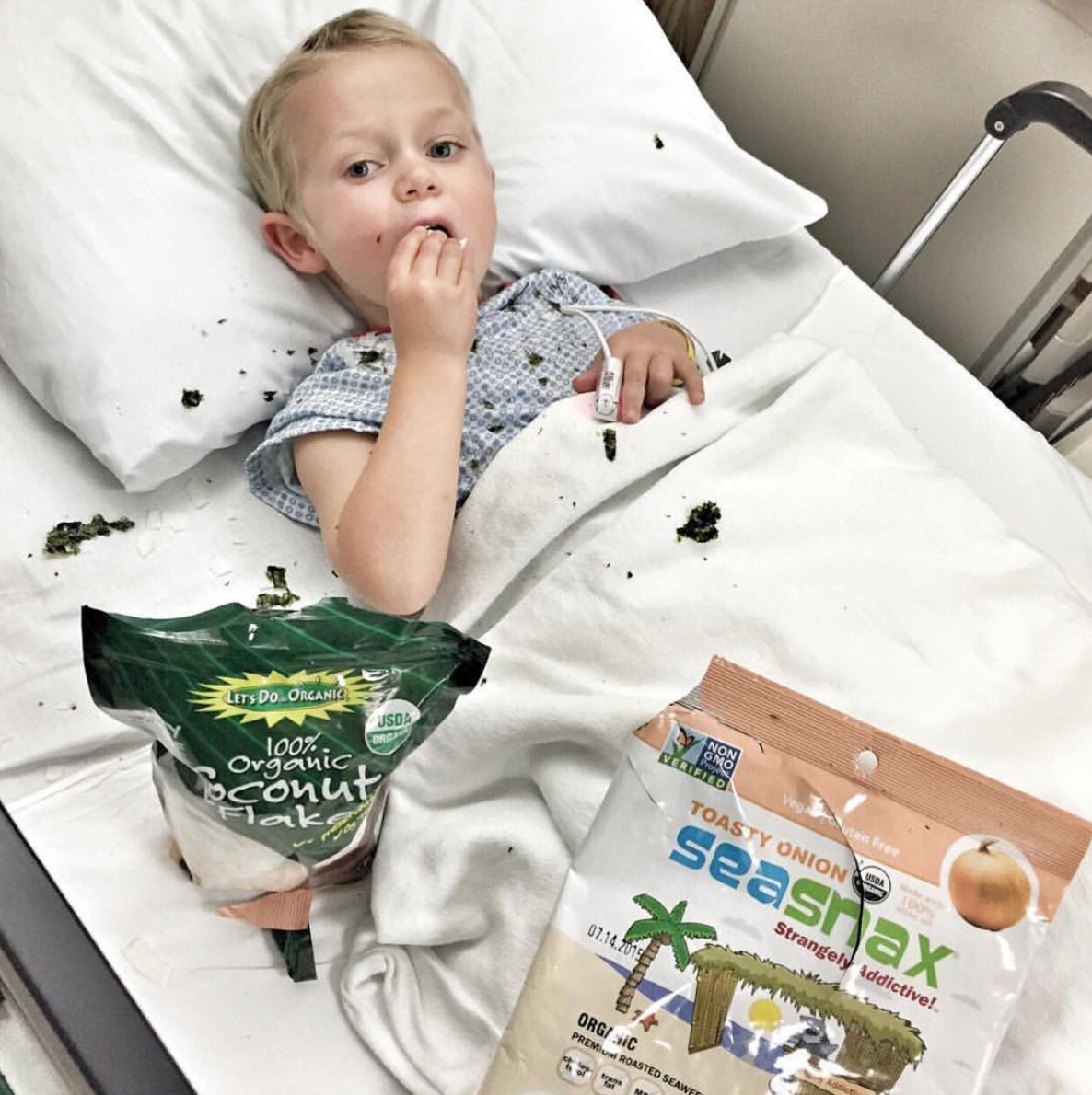 little boy in a hospital gown in a hospital bed looking at the camera eating shredded coconut and seaweed