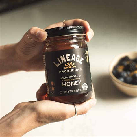 a hand holding a jar of lineage raw honey next to a bowl of berries 